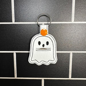 May include: White ghost-shaped keychain with black stitching and a silver key ring. The ghost has a zipper mouth and an orange button on top. The keychain is made of a leather-like material and is a Halloween-themed accessory.