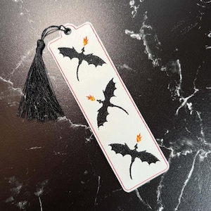 May include: A rectangular white bookmark featuring three black dragon silhouettes, each breathing a small orange and yellow flame. The bookmark has a thin red border and a black tassel attached to the top through a small metal eyelet.