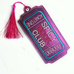 May include: A rectangular purple embroidered bookmark shaped like a ticket with a pink tassel. The text reads ADMIT ONE, SPICY BOOK CLUB, and 690690 in white and red thread. It features a teal border and a white eyelet at the top for the tassel.