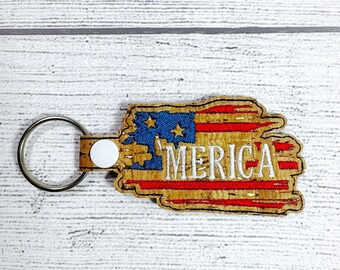 May include: A key chain with a distressed American flag design and the word "MERICA" in white embroidery. The flag is made of cork and has a red, white, and blue color scheme.