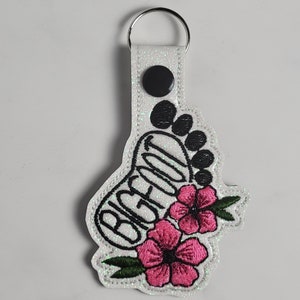 Könnte beinhalten: Weißer Glitzer-Schlüsselanhänger mit einem schwarzen gestickten Fußabdruck und dem Wort "BIGFOOT" in Schwarz. Zwei rosa Blumen mit grünen Blättern sind unter dem Fußabdruck gestickt.