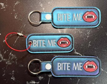 May include: Three rectangular keychains with a blue gradient background and black stitched borders. Each features the white text BITE ME and an embroidered graphic of red lips with white fangs. Two keychains have metal rings, one has a red loop.