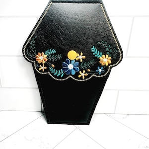 May include: A black faux leather coffin-shaped clutch with a floral embroidery design in shades of orange, blue, green, and yellow. The embroidery features small flowers and leaves.
