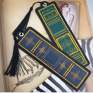 May include: Two rectangular bookmarks with black tassels attached to silver metal eyelets. One bookmark is dark blue with yellow embroidered geometric patterns, and the other is dark green with yellow embroidered floral and grid designs.