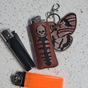 Könnte beinhalten: Ein braunes Leder-Feuerzeugetui mit einem schwarzen Skelett und einem Motten-Design. Das Etui hat einen silbernen Schlüsselring. Neben dem Etui liegen zwei Feuerzeuge, eines schwarz und eines orange.