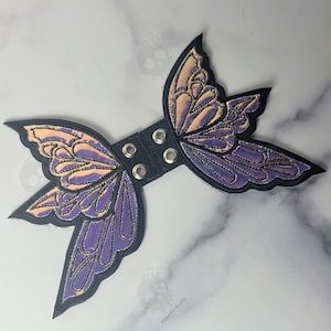 May include: A black, butterfly-shaped accessory with iridescent purple and gold wings. The wings are outlined in black and have intricate embroidered details. The center band is black with silver grommets.