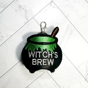May include: Black cauldron-shaped keychain with green bubbling liquid and a wooden spoon. The text "WITCH'S BREW" is embroidered in white. The keychain has a silver-colored clasp.