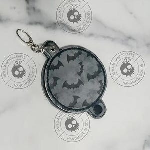 Może przedstawiać: Okrągły brelok w kolorze szarym i czarnym z wzorem nietoperza. Brelok ma metalowy pierścień i czarny guzik. Brelok jest otoczony czterema szarymi i czarnymi okrągłymi logo z tekstem "Asylum Handicrafts Handmade Goods".