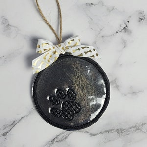 May include: Black and white paw print bauble with a clear plastic window filled with pet fur. The bauble has a white ribbon with gold paw print designs and a brown twine hanger.