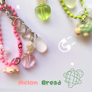 Op de afbeelding: Twee decoratieve telefoonbedels met diverse kralen en bedels. Eén is roze met een hart- en sterbedel, de andere is groen met een hart-, ster- en schildpadbedel. De bedels hebben een lichtgroen koord.