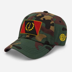 May include: Camouflage baseball cap with a black, red, and gold patch featuring a spear and wreath design. The cap also has a yellow "FBA" logo on the side. The hat is made of a green, brown, and black camouflage pattern.