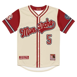 May include: Cream baseball jersey with red sleeves and trim. The jersey displays the word "Monarchs" in a script font, the number 5, and several patches and logos. Includes text "1962 Hall of Fame Jackie Robinson #5".