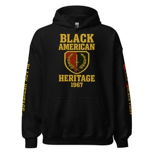 Puede incluir: Sudadera con capucha negra con texto dorado y un diseño de escudo. El texto "BLACK AMERICAN HERITAGE 1967" está impreso en la parte delantera. La manga izquierda tiene "BLACK HERITAGE" en dorado. La manga derecha tiene "BLACK FIRST" en dorado.