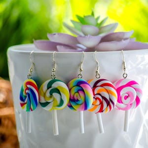 May include: A collection of five colorful lollipop earrings. Each earring features a swirled design in various colors, including blue, green, yellow, orange, red, and pink, attached to a white stick. The earrings are displayed against a white background.