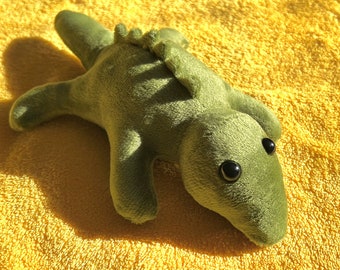 May include: A green plush alligator toy with black eyes. The toy is lying on a yellow towel.