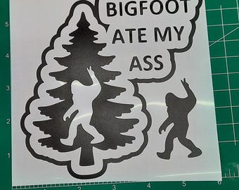 May include: A white sticker with a black graphic of a Bigfoot figure and a tree. The text "BIGFOOT ATE MY ASS" is in a speech bubble. The sticker is on a green cutting mat.