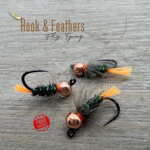 May include: Three fly fishing lures featuring black hooks, metallic copper beads, dark green iridescent bodies, and bright orange synthetic fiber tails. The text Hook & Feathers Fly Tying is visible at the top of the image.