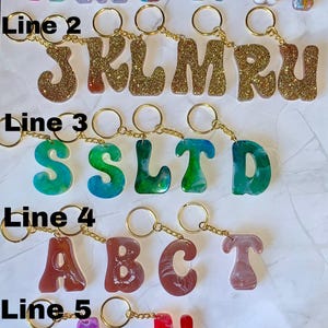Può includere: Una collezione di portachiavi con lettere dell'alfabeto in vari colori e design. Ogni lettera è attaccata a una catena e un portachiavi dorati. Le lettere presentano glitter, motivi floreali ed effetti marmorizzati, creando un accessorio personalizzato.