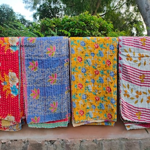 May include: Four kantha quilts hanging on a wall. The quilts are made of recycled saris and have different floral patterns. The quilts are in red, blue, yellow, and white with red stripes.