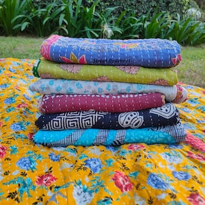 May include: A stack of six colorful kantha quilts, folded and arranged on a yellow floral kantha quilt. The quilts are made of cotton and have a variety of patterns, including floral, geometric, and paisley. The text "Street Bedsheet" is visible on the top quilt.
