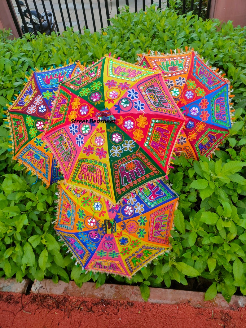 Puede incluir: Un conjunto de cuatro paraguas coloridos con bordados intrincados y adornos de espejo. Los paraguas est&aacute;n decorados con motivos florales y elefantes. Los paraguas est&aacute;n apilados, con el paraguas superior parcialmente abierto.