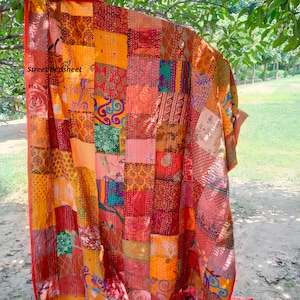 Könnte beinhalten: Eine farbenfrohe Patchwork-Decke mit verschiedenen Mustern und Farben, darunter Rot, Orange, Gelb, Grün und Blau. Die Decke ist aus recycelten Saris gefertigt und handgenäht. Die Decke hängt an einem Baumzweig.