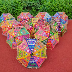 Puede incluir: Un grupo de ocho paraguas bordados de colores con ribetes de flecos. Los paraguas est&aacute;n dispuestos en c&iacute;rculo sobre una superficie roja. Los paraguas est&aacute;n decorados con varios patrones y colores, incluyendo rosa, azul, verde, amarillo y naranja. Los paraguas est&aacute;n hechos de tela y tienen un mango de madera.
