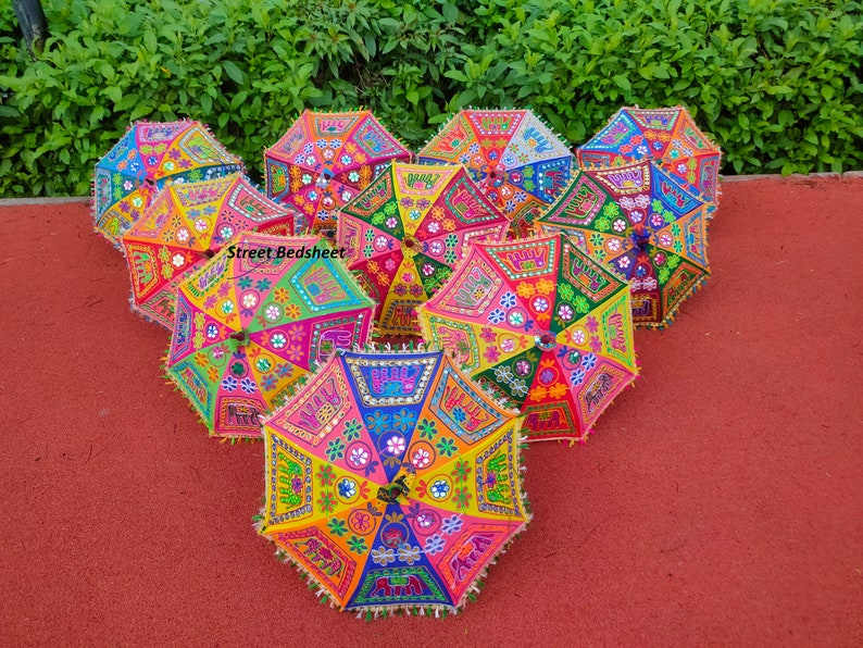 Puede incluir: Un grupo de ocho paraguas bordados de colores con ribetes de flecos. Los paraguas est&aacute;n dispuestos en c&iacute;rculo sobre una superficie roja. Los paraguas est&aacute;n decorados con varios patrones y colores, incluyendo rosa, azul, verde, amarillo y naranja. Los paraguas est&aacute;n hechos de tela y tienen un mango de madera.