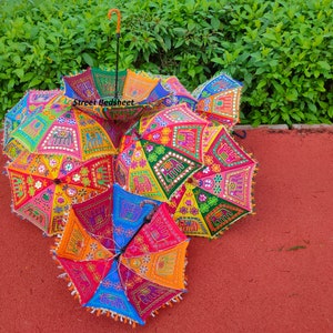Puede incluir: Un grupo de seis paraguas coloridos con bordados intrincados y adornos de espejo. Los paraguas est&aacute;n hechos de tela y tienen un dise&ntilde;o tradicional indio. Los paraguas est&aacute;n dispuestos en una pila sobre una superficie roja.