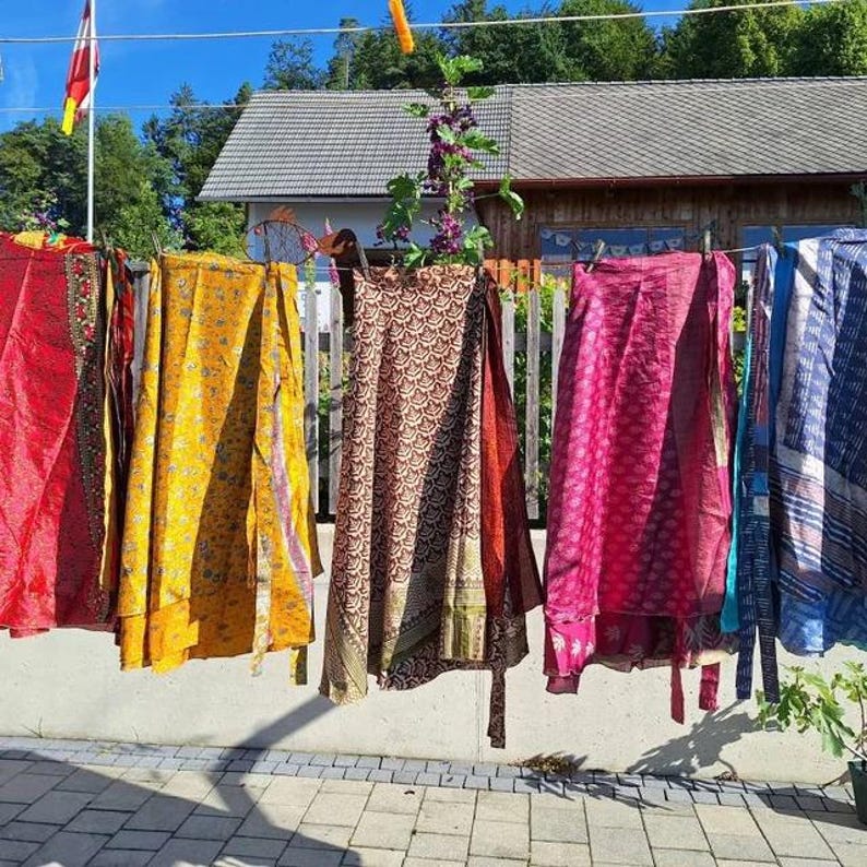 May include: Five wrap skirts in various colors and patterns, including red, yellow, brown, pink, and blue, are hanging on a clothesline. Each skirt features a distinct print and is suspended by clips, displaying a flowing, lightweight fabric construction.