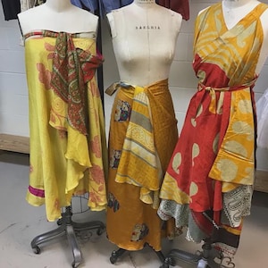 May include: Three dress forms display wrap-style garments. The left form wears a yellow dress with red floral patterns. The center form wears a mustard yellow wrap skirt with elephant motifs. The right form wears a red and yellow paisley dress.
