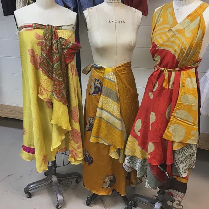 May include: Three dress forms display wrap-style garments. The left form wears a yellow dress with red floral patterns. The center form wears a mustard yellow wrap skirt with elephant motifs. The right form wears a red and yellow paisley dress.