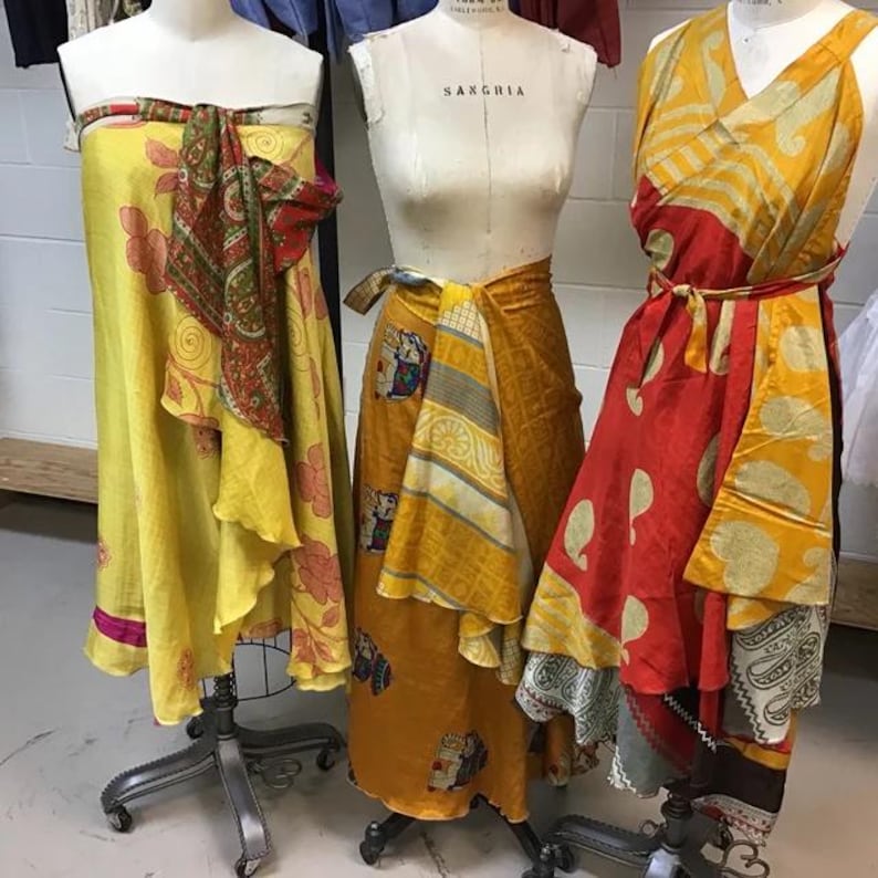 May include: Three dress forms display wrap skirts in vibrant colors. The left skirt is bright yellow with red floral patterns. The center skirt is mustard yellow with elephant motifs. The right skirt features a red and yellow paisley print with a tie waist.