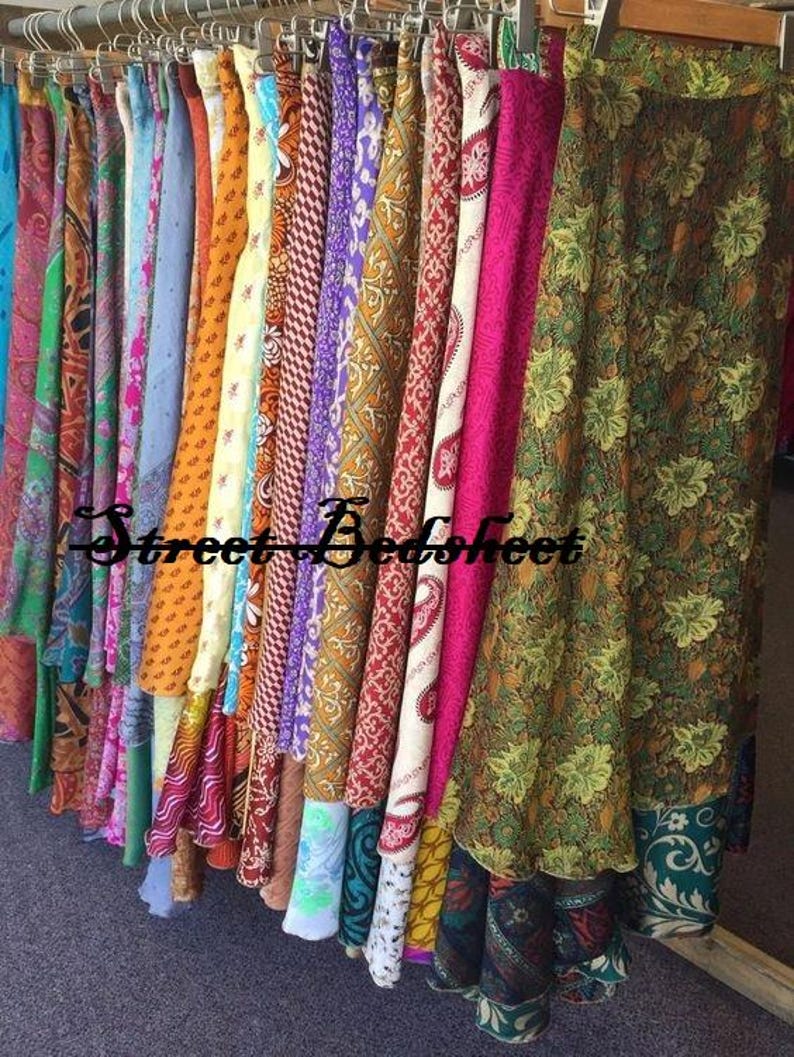 May include: A collection of long skirts hanging on a rack, featuring a variety of colorful fabrics with diverse patterns including floral, paisley, and geometric designs. The text Street Bedsheet is overlaid across the center of the hanging garments.