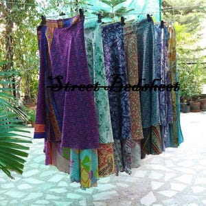 May include: A collection of long wrap skirts hanging on a clothesline, featuring various vibrant colors and intricate floral and geometric patterns. The text Street Bedsheet is overlaid in black across the center of the hanging fabric garments.