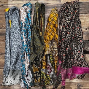 May include: Five long, rectangular fabric wraps displayed side-by-side. The first is grey with a paisley pattern, the second is blue and white with floral motifs, the third is olive green with swirls, the fourth is yellow plaid, and the fifth is black with floral prints and a pink hem.