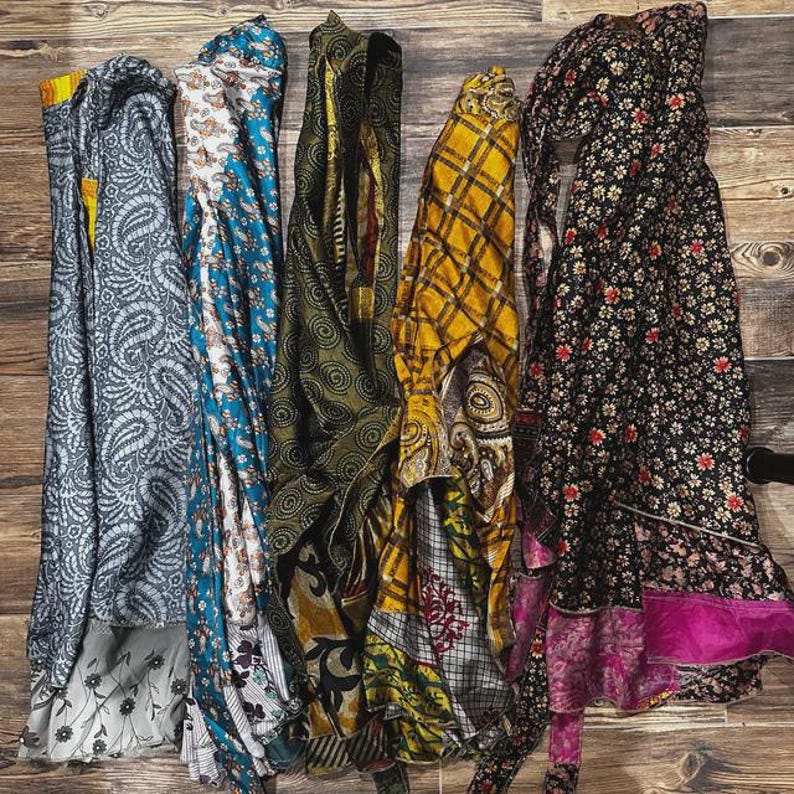 May include: Five long, rectangular fabric wraps displayed side-by-side. The first is grey with a paisley pattern, the second is blue and white with floral motifs, the third is olive green with swirls, the fourth is yellow plaid, and the fifth is black with floral prints and a pink hem.