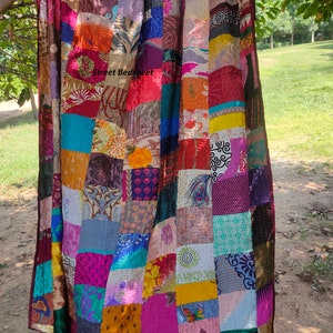 May include: A colorful patchwork quilt made from repurposed saris. The quilt is made up of squares of different colors and patterns, including floral, geometric, and paisley designs. The quilt is hanging from a tree branch and has the text "Street Bedsheet" printed on it.