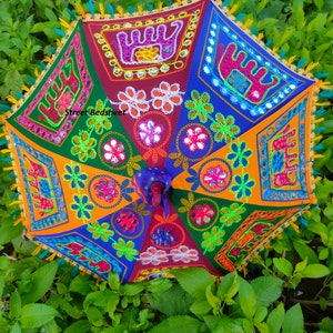 Puede incluir: Un paraguas colorido y multi-patr&oacute;n con detalles bordados y acentos de espejo. El paraguas est&aacute; hecho de tela y tiene un mango de madera. El paraguas est&aacute; rodeado de follaje verde.