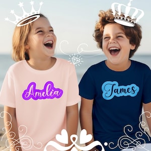 May include: Two children wearing t-shirts with names. One child wears a light pink shirt with the name "Amelia" in purple script. The other child wears a navy blue shirt with the name "James" in light blue script. Both children are smiling and wearing crowns.