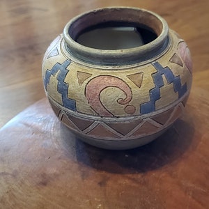 May include: A small, round, handcrafted ceramic pot with a beige base and geometric patterns in blue, red, and brown. The pot has a wide opening and a slightly textured surface, showcasing a traditional design.
