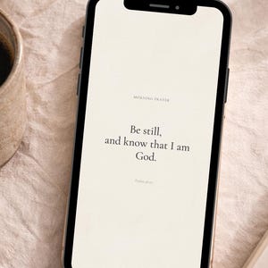 May include: A smartphone with a white screen displaying the text "Be still, and know that I am God." is positioned next to a cup of coffee and a notebook with a pen. The words "MORNING PRAYER" and "PHONE LOCK SCREEN" are at the top.