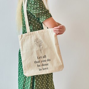 Peut inclure: Un sac fourre-tout en toile blanche avec du texte noir qui dit "Let all that you do be done in love". Le sac présente également un dessin au trait simple d'une fleur.