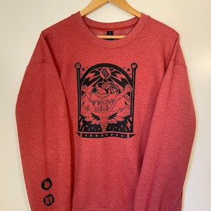 May include: Red sweatshirt with a black graphic design featuring a stylized creature with outstretched arms, set within a rectangular frame. The sweatshirt has a crew neck and long sleeves, with two black symbols on the left sleeve.