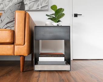 May include: A black metal end table with a shelf and a drawer. The table has a concrete base and is holding a stack of books. The table is next to a brown leather sofa.