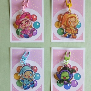 Puede incluir: Cuatro llaveros de acrílico de colores con personajes de estilo anime. Cada dije tiene un diseño de personaje diferente con colores vibrantes, adjunto a una tarjeta rosa con un diseño de burbujas y un cierre de metal.
