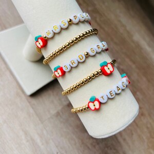 May include: Four gold beaded bracelets with white letter beads spelling "MRS. PLUM", "3RD GRADE", and "TEACHER" with red apple charms.
