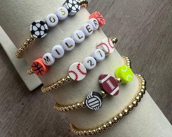 May include: Four gold beaded bracelets with different sports-themed charms. The charms include a soccer ball, basketball, baseball, football, volleyball, and tennis ball. The bracelets also have letter beads that spell out "MILLER" and the numbers "05" and "21".