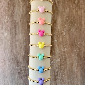 May include: A collection of beaded bracelets in various colors, each featuring a Mickey Mouse-shaped charm. The bracelets are displayed on a white cylindrical stand against a brown background. Colors include cream, pink, coral, fuchsia, yellow, teal, blue, and purple.