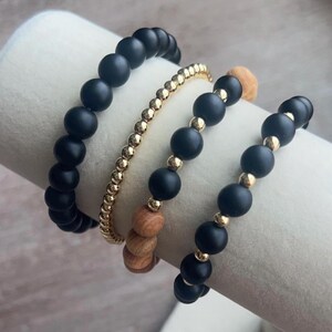 May include: A collection of beaded bracelets in various styles. The bracelets feature black matte beads, gold-colored beads, and wooden beads. The bracelets are displayed on a white cylindrical surface.